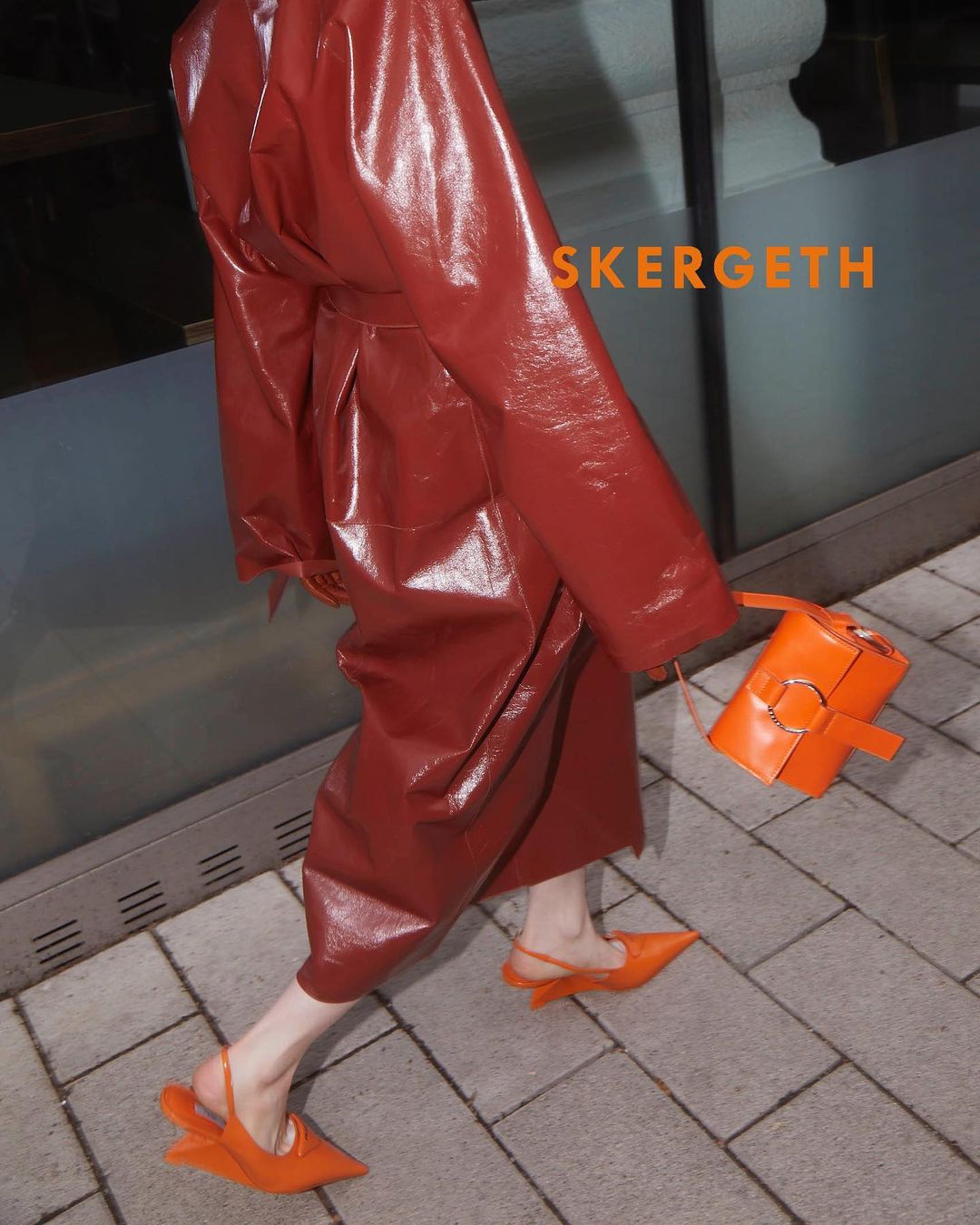 Fashionable woman walking in orange slingback shoes and a matching handbag, wearing a shiny copper trench coat on a sidewalk.