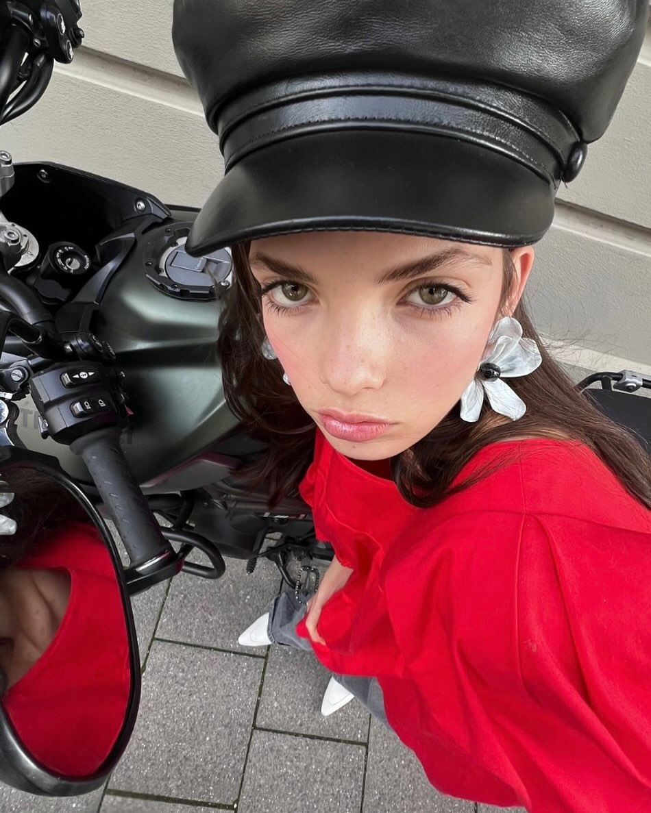Woman in a black cap and red shirt stands next to a motorcycle on a city street with light-colored paving stones.