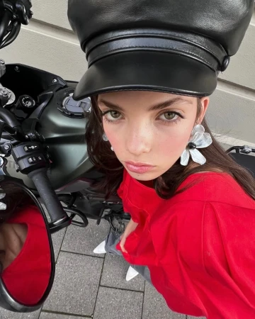 Woman in a black cap and red shirt stands next to a motorcycle on a city street with light-colored paving stones.