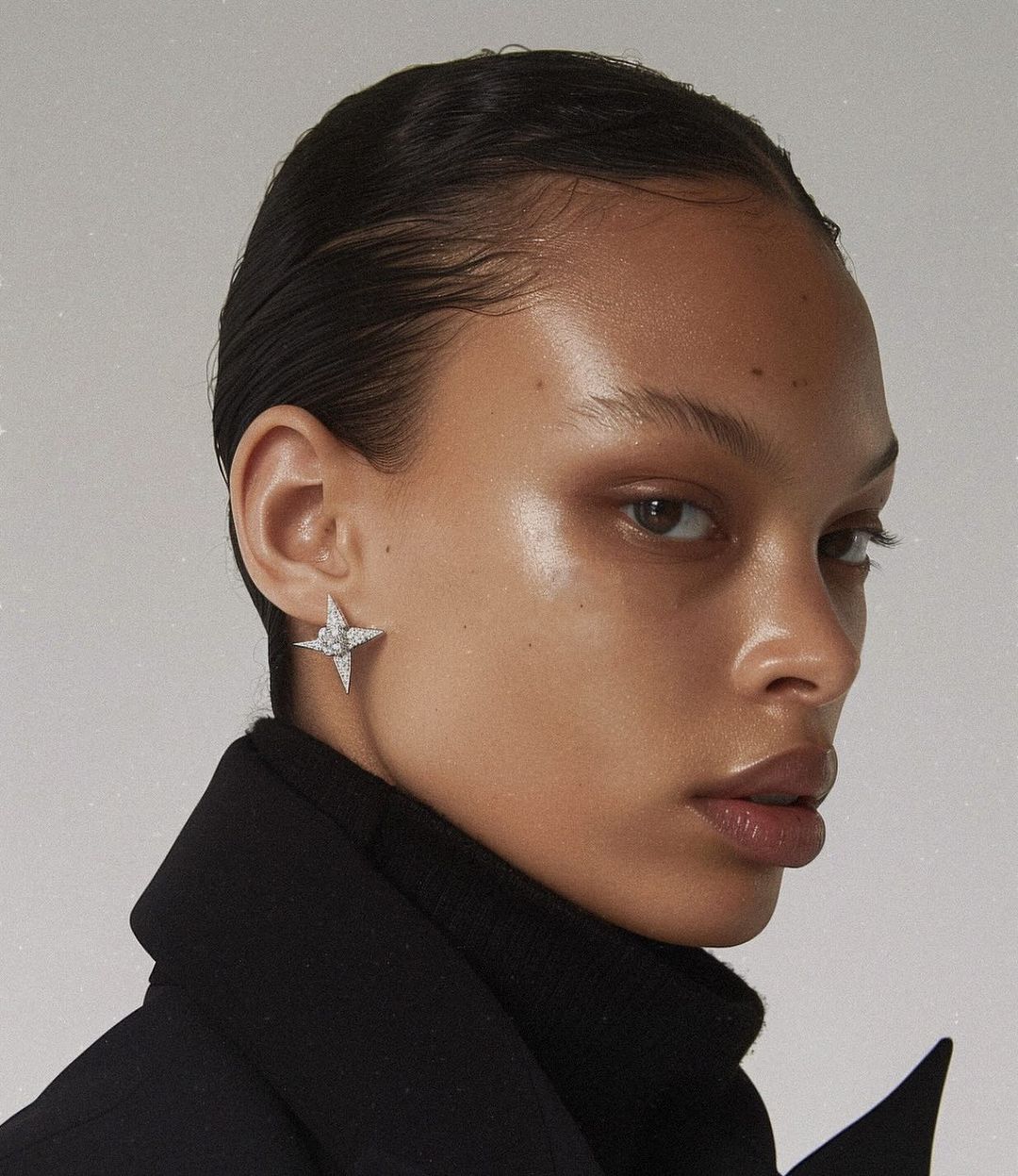 Portrait of a woman with dark hair wearing diamond star earrings and a black coat, posing against a white background in studio lighting.