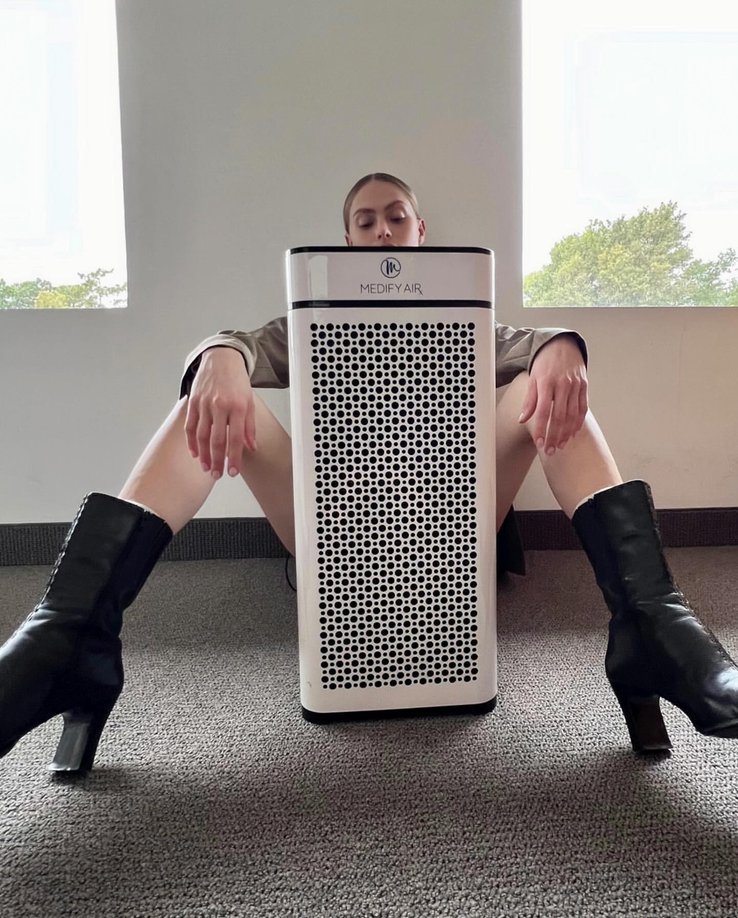 Adult female poses indoors with air purifier in a studio-like setting, showcasing a modern lifestyle product near a window with natural light.
