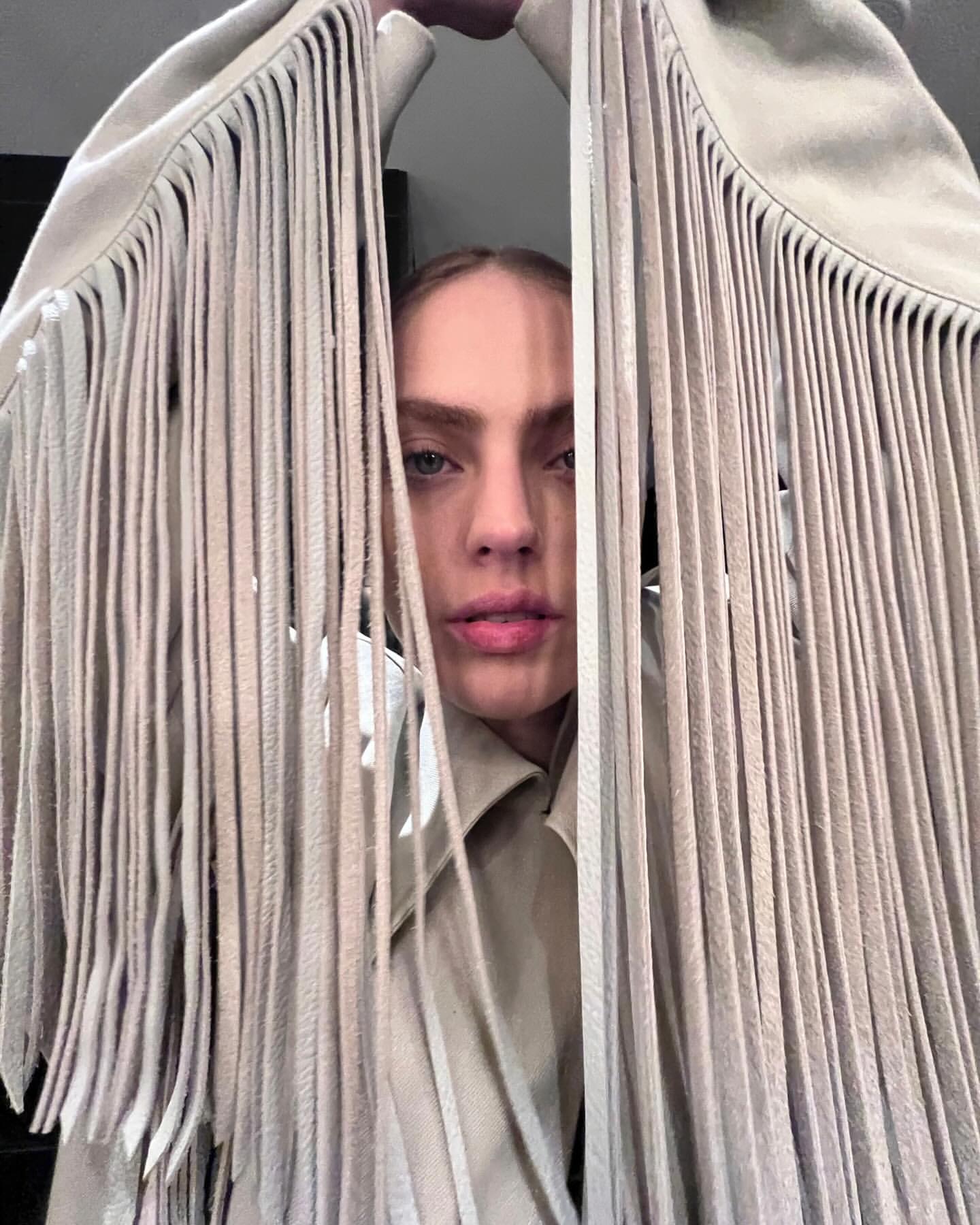 Portrait of a woman framed by a beige jacket with fringe hanging around her face, creating a peek-a-boo effect in an indoor setting.