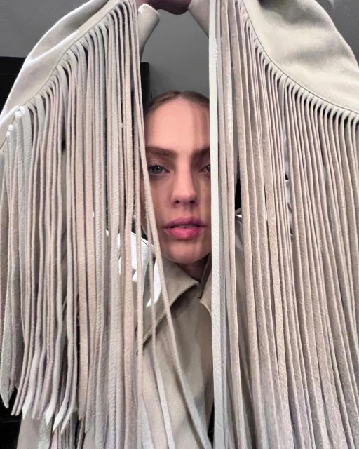 Portrait of a woman framed by a beige jacket with fringe hanging around her face, creating a peek-a-boo effect in an indoor setting.
