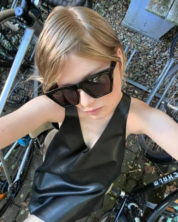 Close-up selfie of a blonde woman wearing black sunglasses and a black leather top, seated near bicycles.
