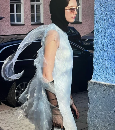 Model in translucent blue dress and black hood standing on city street by parked cars