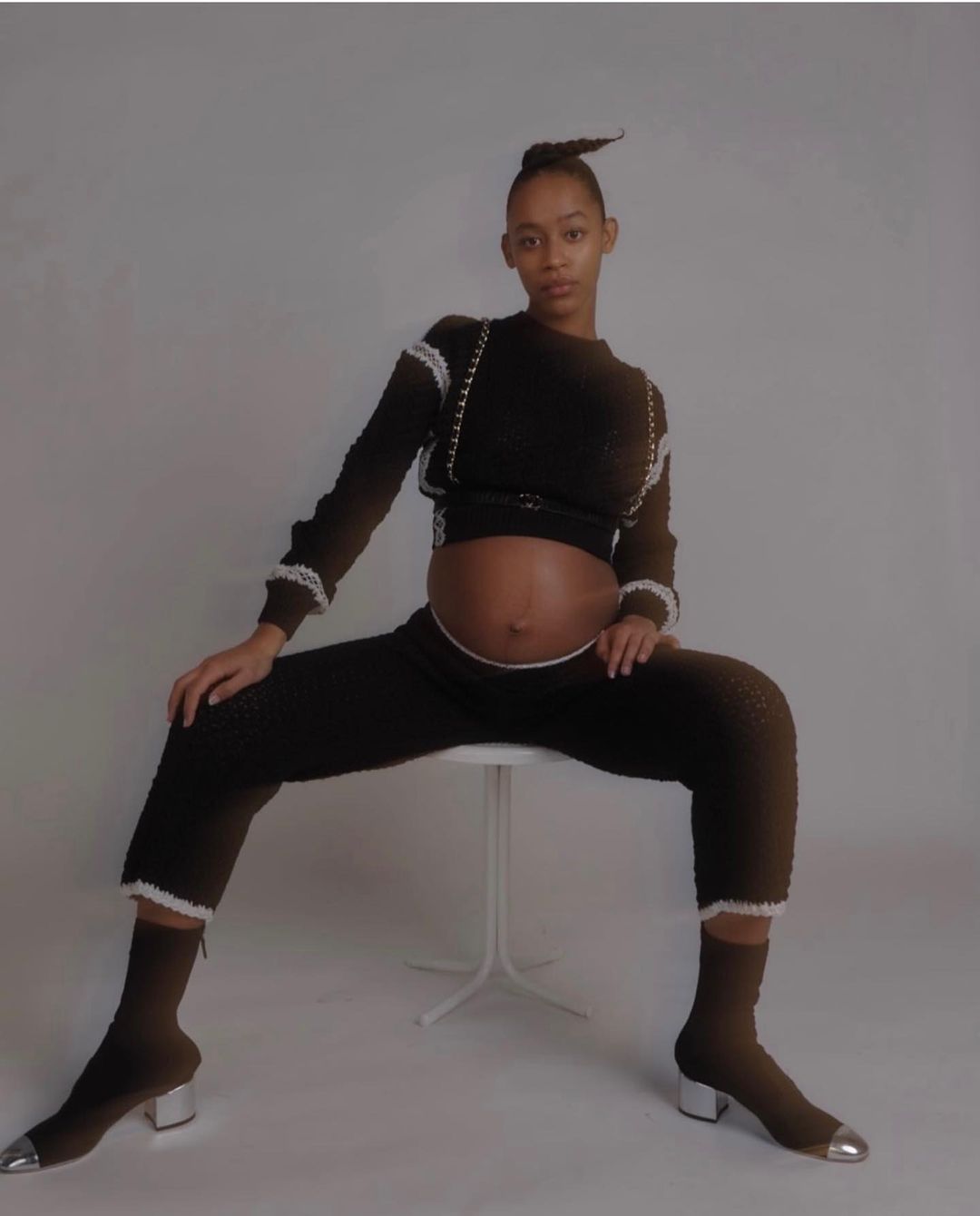 Pregnant African-American woman in a dark knitwear outfit sitting on a white stool in a light gray studio setting looking at the camera.