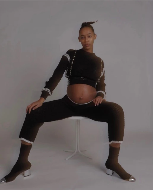 Pregnant African-American woman in a dark knitwear outfit sitting on a white stool in a light gray studio setting looking at the camera.
