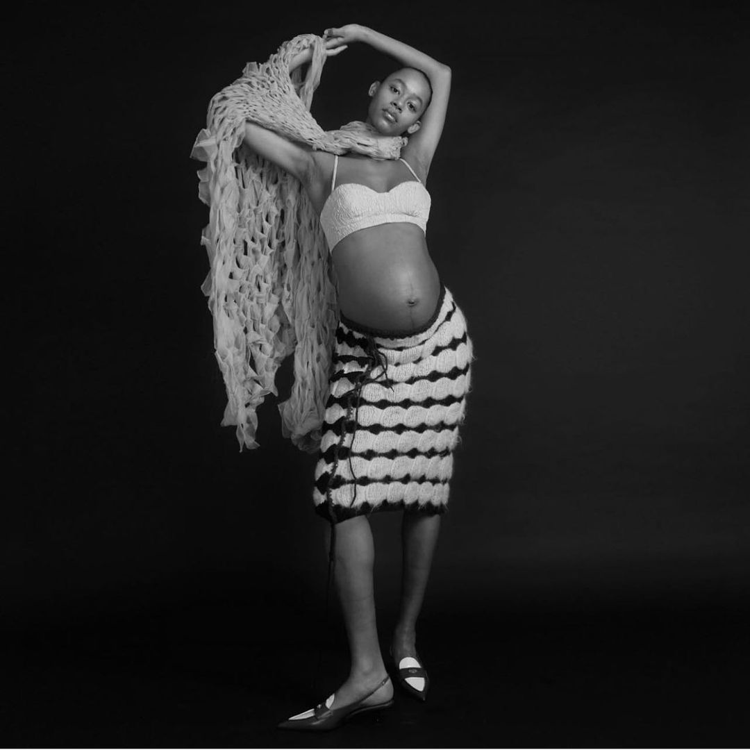 Striking studio portrait of a pregnant African-American woman in knitted crop top and skirt with draped scarf against a dark background.
