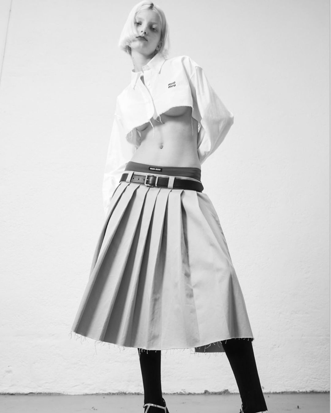 A low-angle, black-and-white fashion shot of a slender adult female model with short blonde hair wearing a white crop top and pleated skirt.