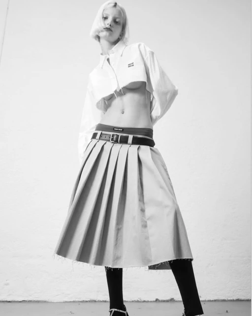 A low-angle, black-and-white fashion shot of a slender adult female model with short blonde hair wearing a white crop top and pleated skirt.