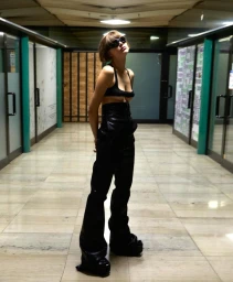 Model wearing dark sunglasses, a black top, and black trousers posing inside an interior hallway with tiled floors.