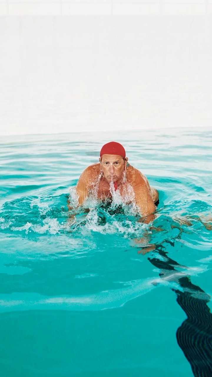 Adult male swimmer in red swim cap swims breaststroke in indoor pool, water splashing around him, white tiled wall visible.