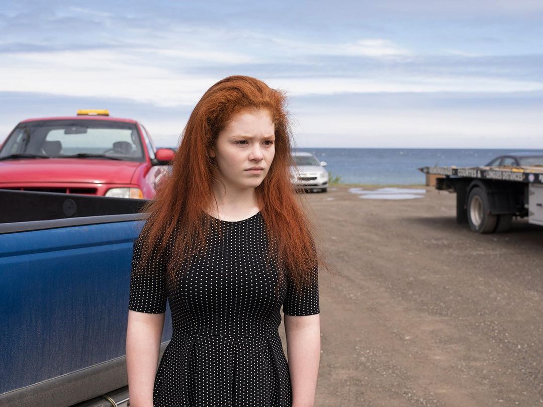 Young woman with red hair stands near pickup trucks and the ocean in a medium shot on a sunny day with bright lighting.
