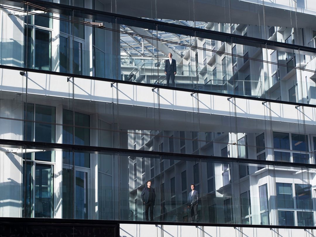 Three men in business suits are visible through the glass exterior of a modern office building on separate levels, reflecting the city.