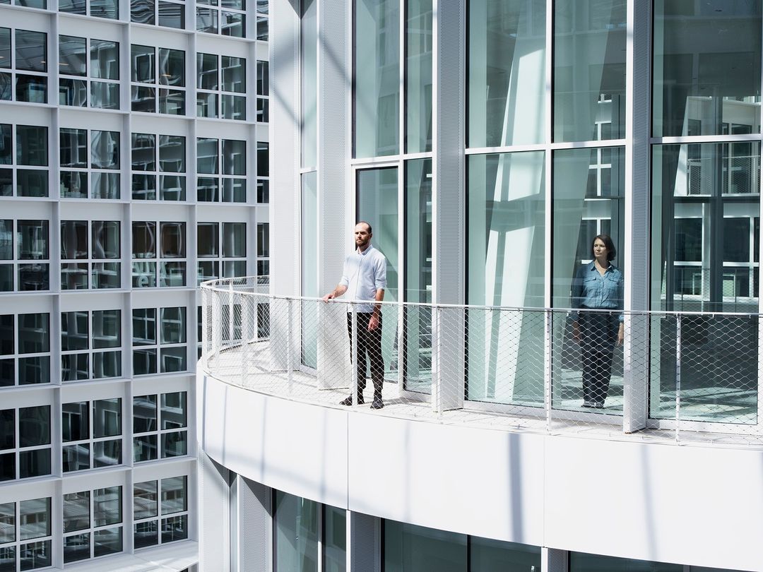 Two people stand on a balcony with a metal railing, in front of a building with glass windows, on a bright day, in an urban setting.