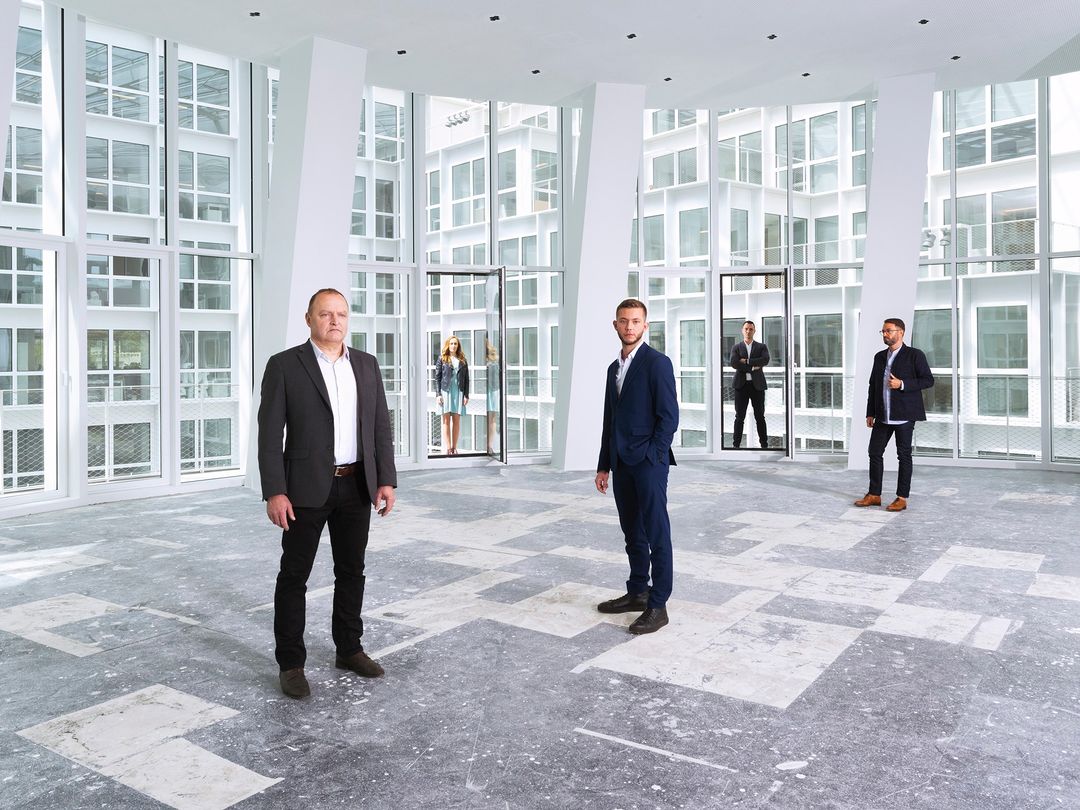 Group of people wearing suits stand inside a modern architectural building with large windows, concrete floors, and white structural beams.