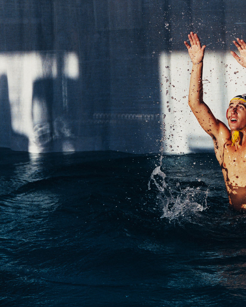 Man in swimming pool with arms raised, droplets frozen in air, wearing a headband, indoor pool with blue-gray walls, bright natural light.
