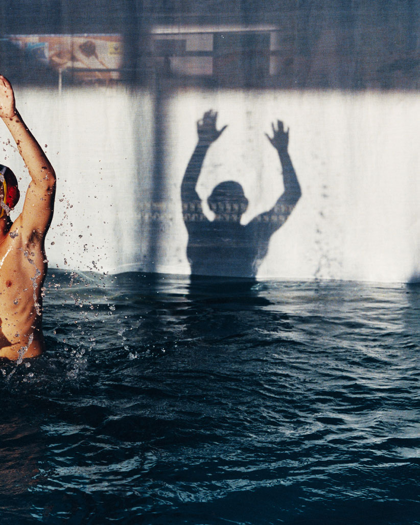 Water polo player in a pool with arms raised and a shadow figure behind. Dark blue water is textured with sunlight.