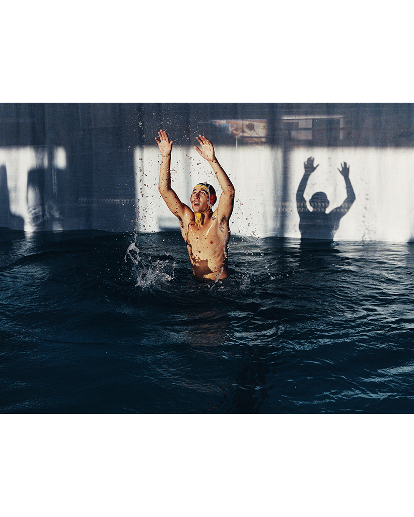 Man in a swimming pool with his arms raised, capturing motion and water splash; a blurred silhouette is seen against tiled wall.