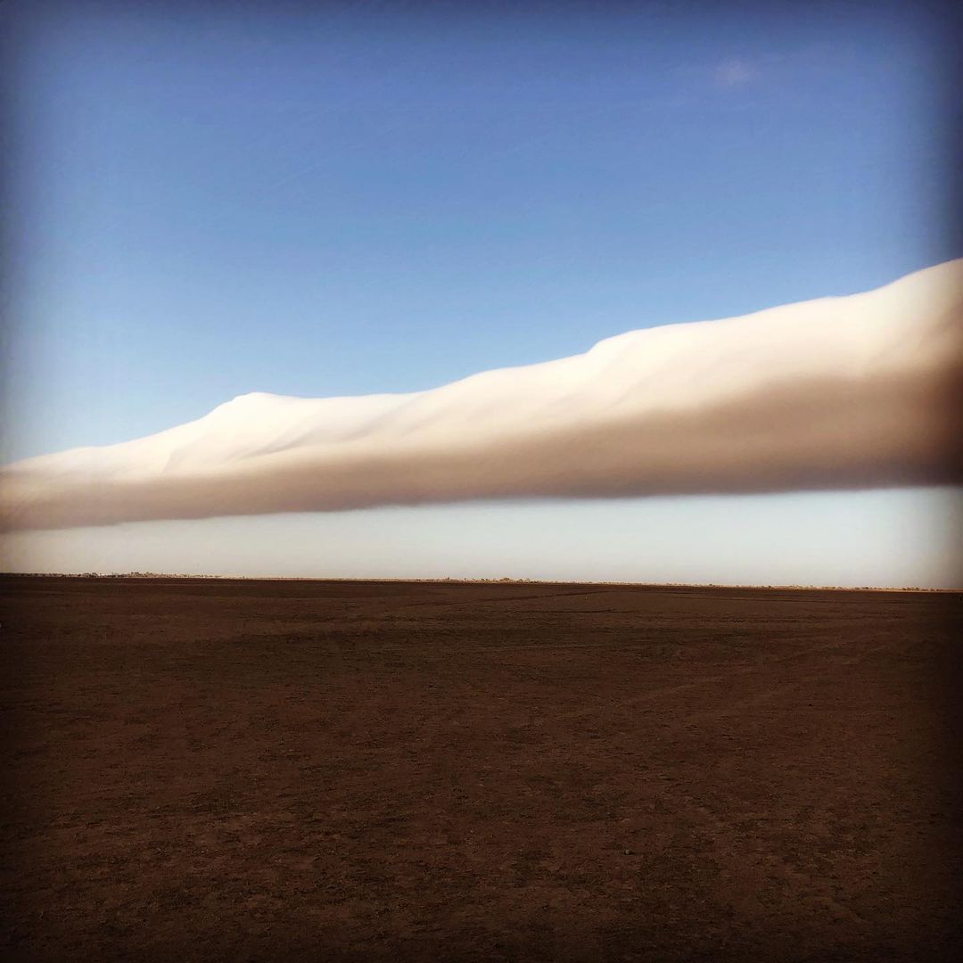 Scenic landscape featuring a clear blue sky, a long, thick band of clouds, and a vast, flat expanse of brown field stretching to the horizon.