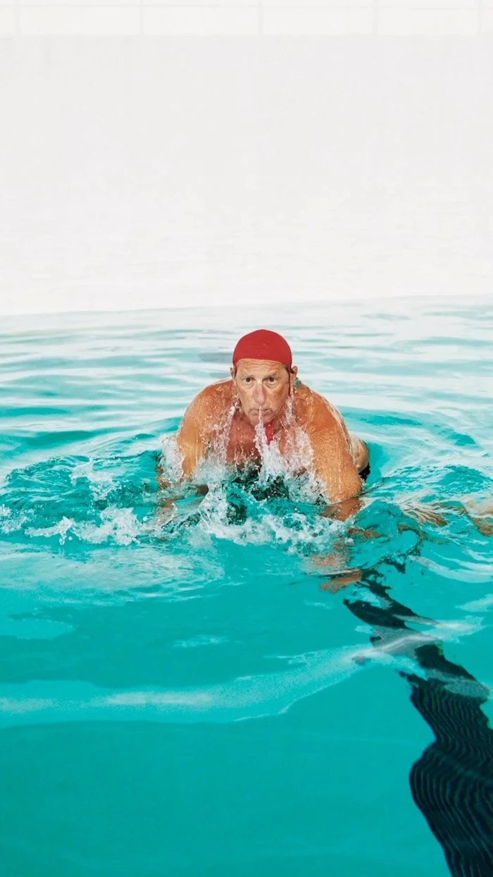 Elderly man wearing a red swim cap swimming breaststroke in a bright turquoise outdoor pool.