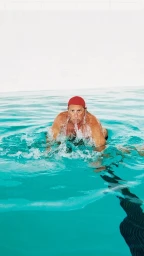 Elderly man wearing a red swim cap swimming breaststroke in a bright turquoise outdoor pool.