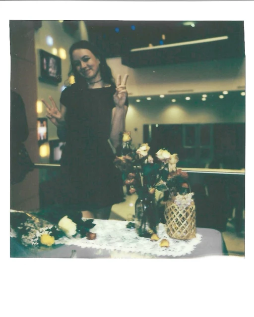 Young woman in a dark dress flashes peace signs next to flower arrangements at an indoor event with mirrored walls and warm lighting.