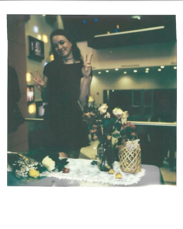 Young woman in a dark dress flashes peace signs next to flower arrangements at an indoor event with mirrored walls and warm lighting.
