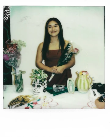 Portrait of a young woman in a red dress holding a single rose. The photo is taken indoors with flowers and objects on a table.
