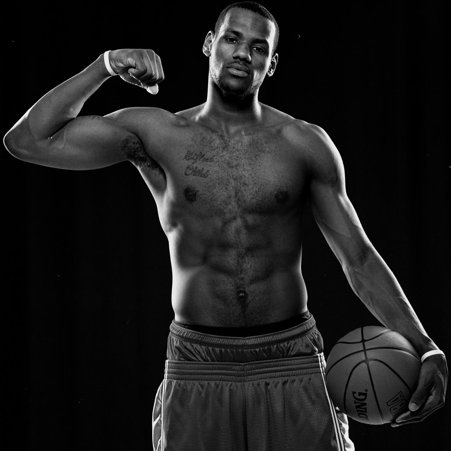 Black-and-white portrait of a muscular Black adult male athlete with a basketball, flexing his arm in a dark photo studio.