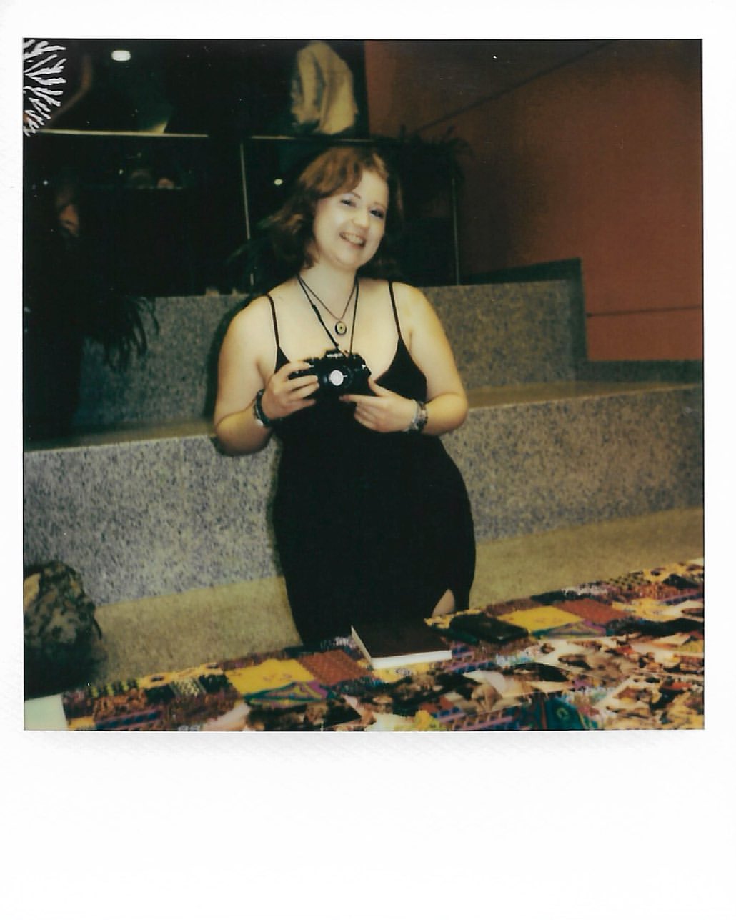 Smiling woman in black dress poses with vintage camera at a table covered in colorful textiles indoors. Bright lighting.