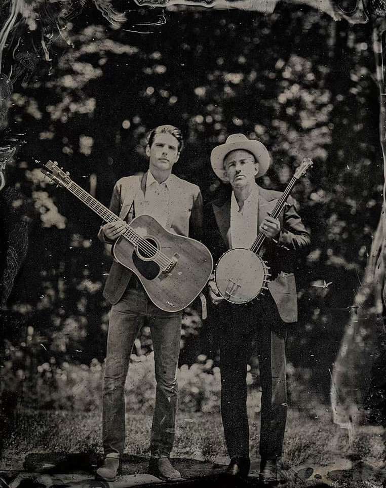 Two men outdoors, one holding an acoustic guitar and the other a banjo. They are standing in front of a background of trees.