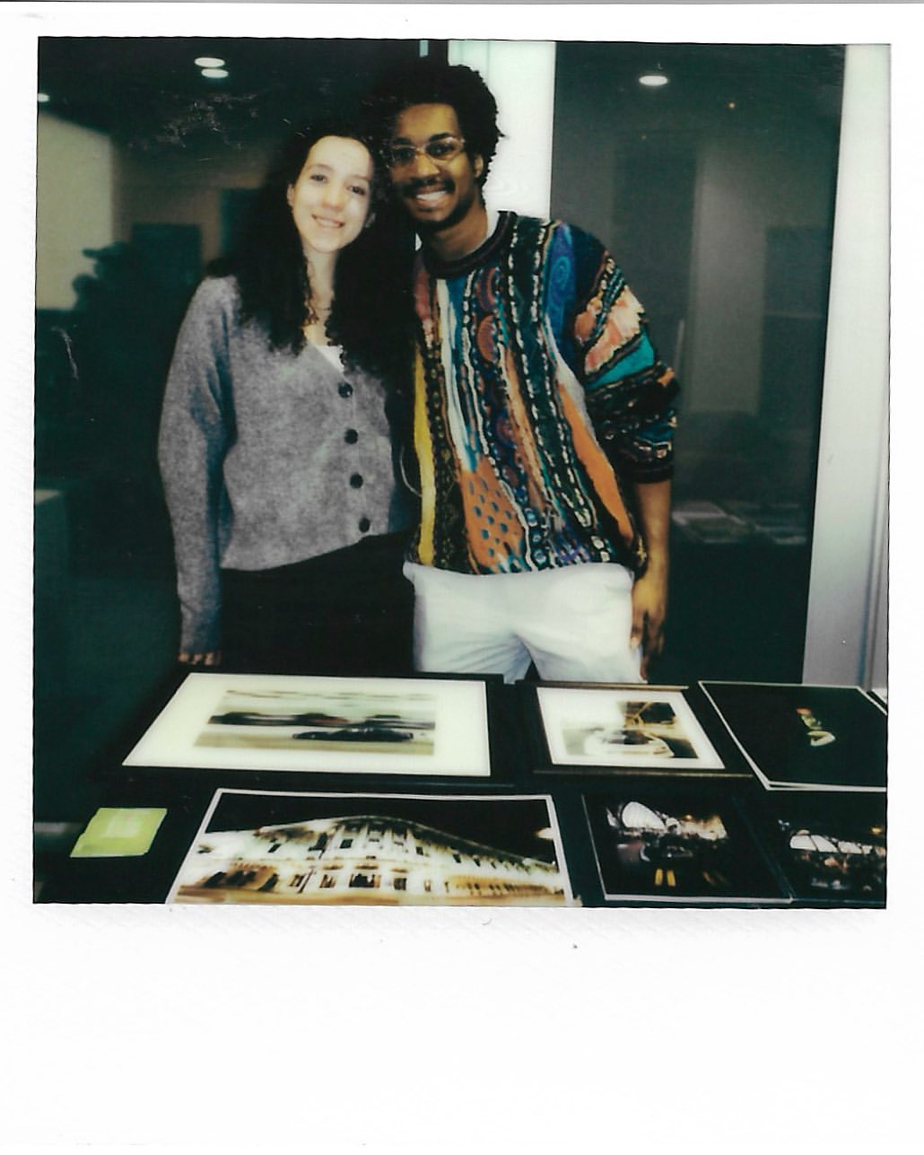 Two smiling people stand behind a table displaying framed images. A light gray cardigan and a colorful sweater are visible.