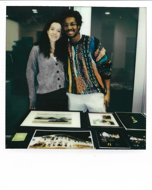 Two smiling people stand behind a table displaying framed images. A light gray cardigan and a colorful sweater are visible.