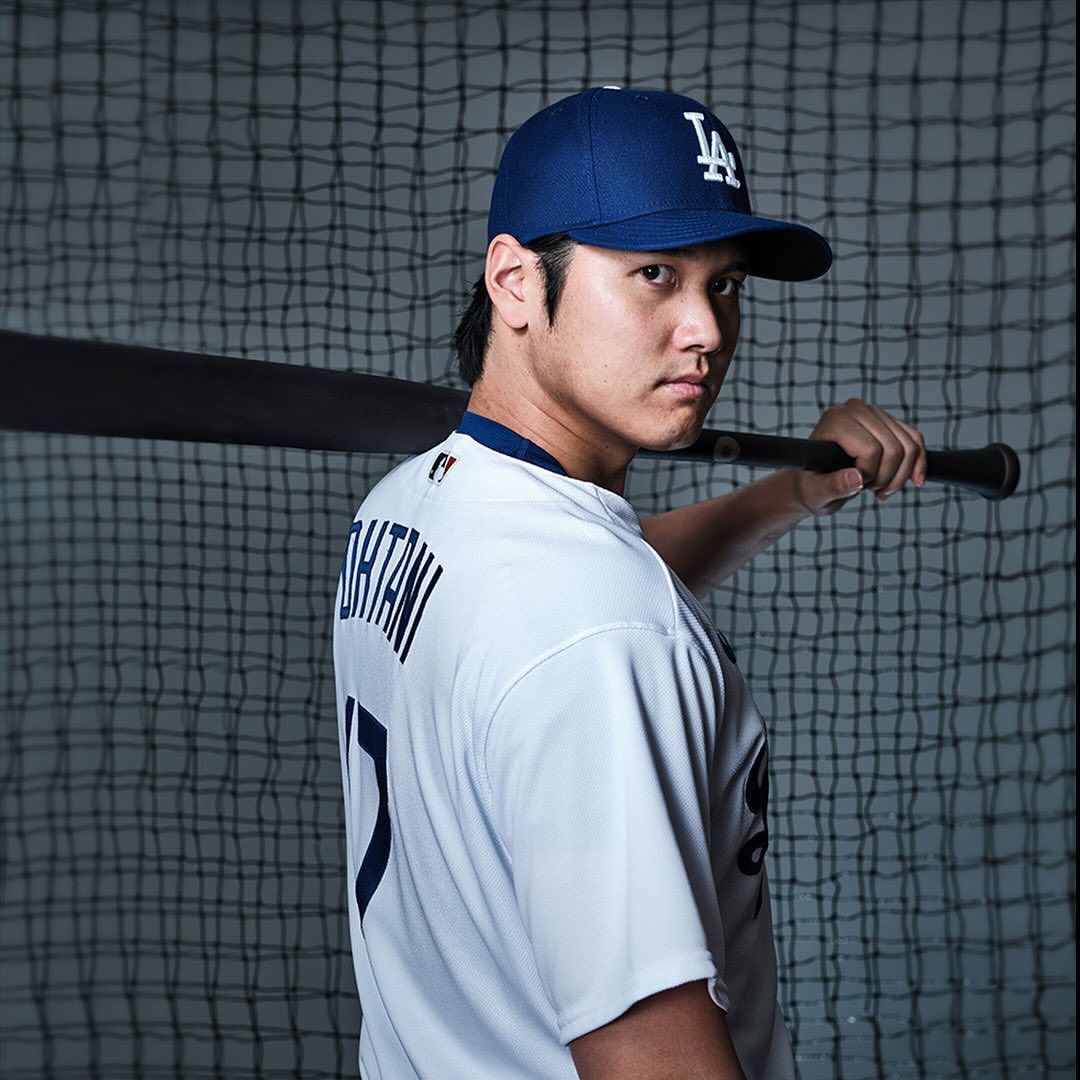 Shohei Ohtani in a white Los Angeles Dodgers baseball jersey and blue cap, holding a bat over his shoulder and looking at the camera.