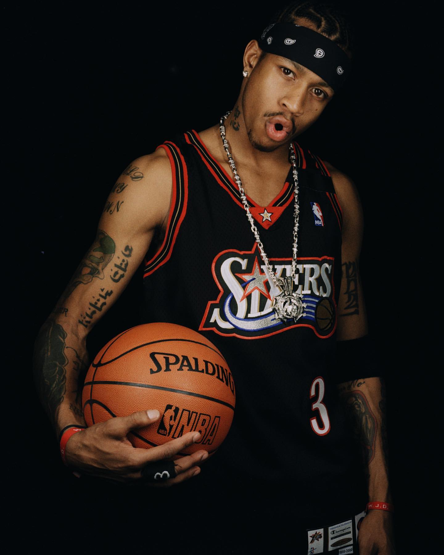 Allen Iverson poses in a 76ers jersey with a basketball against a dark backdrop, showcasing sports and celebrity appeal.