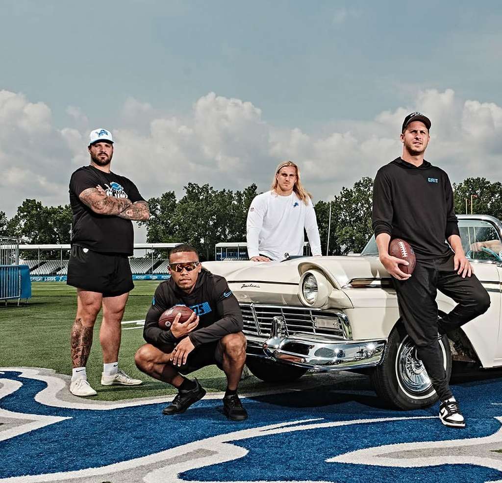 Four football players pose beside a vintage white car on a blue field with the Detroit Lions logo. Clear sky in the background.