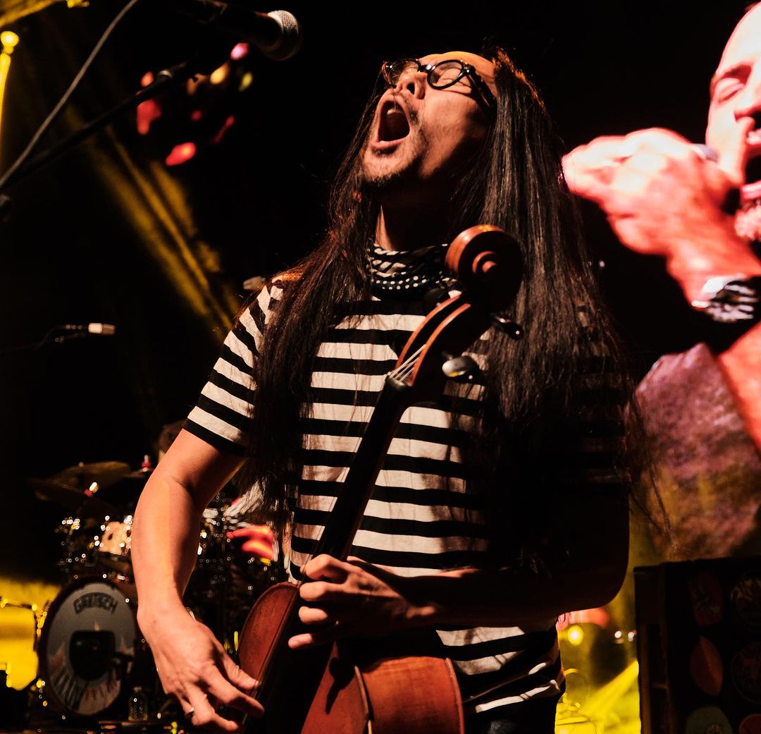 A musician sings enthusiastically while playing a cello on stage at a music venue. The lighting is dim with bright yellow accents.