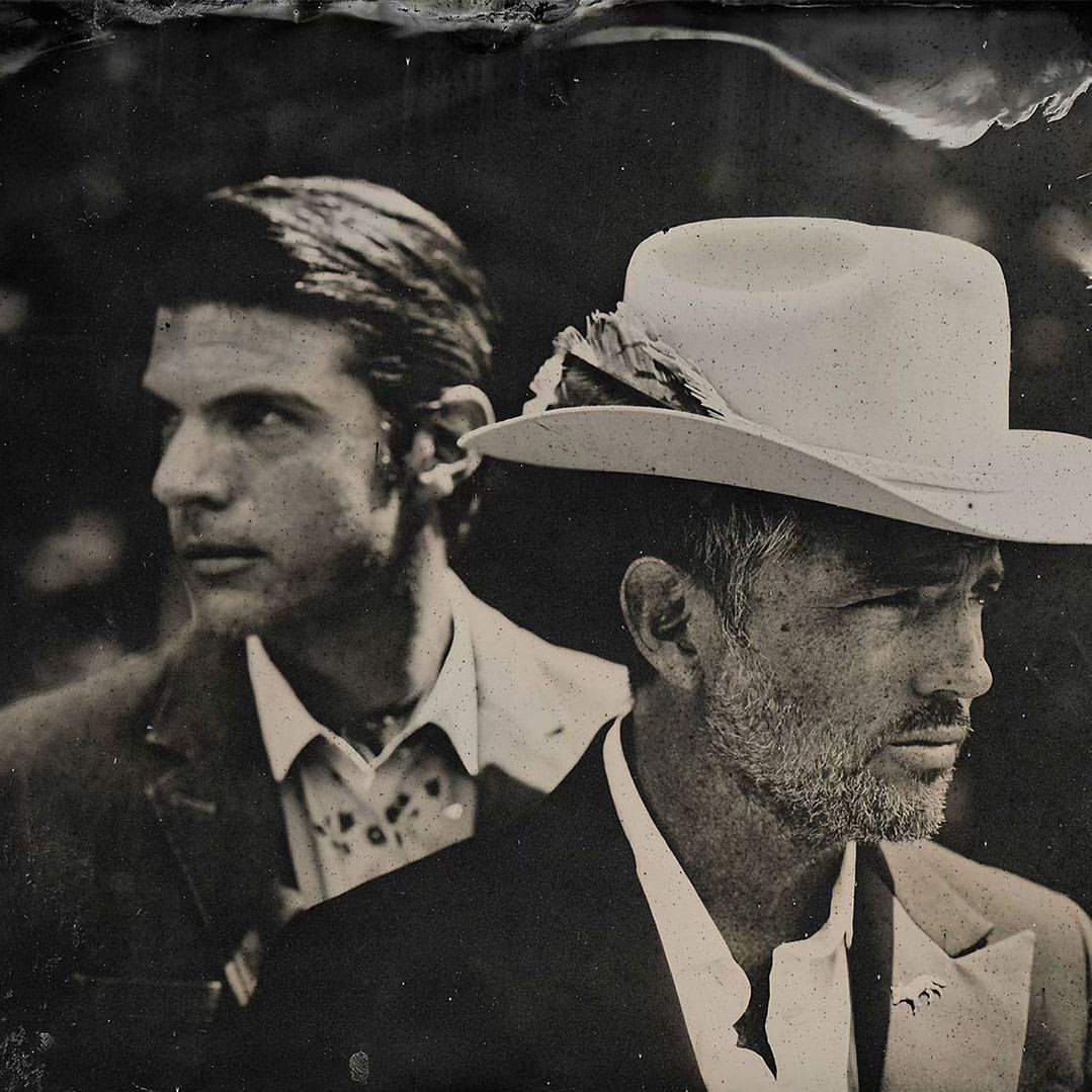 Black and white portrait of two adult men in suits, one wearing a cowboy hat with a feather, looking off to the side.