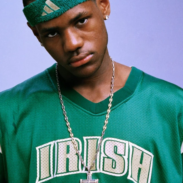 Portrait of an adult male wearing a green sports jersey and headband with a silver chain against a gradient purple background.