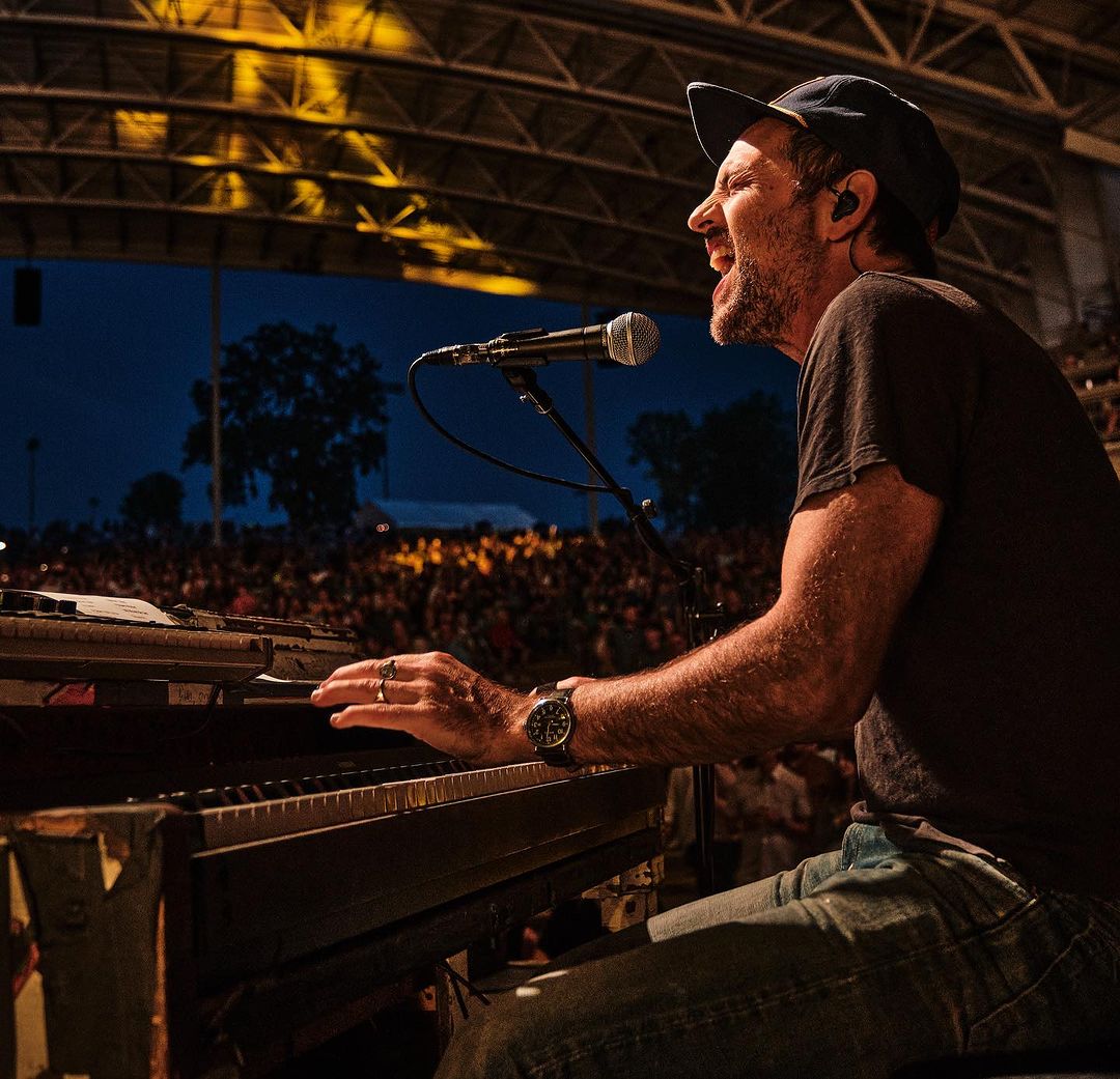 Man passionately singing and playing keyboard at outdoor concert in dark t-shirt, jeans, and ball cap in front of a crowd.