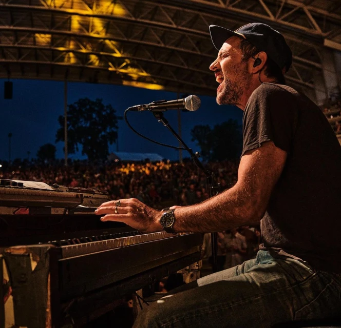 Man passionately singing and playing keyboard at outdoor concert in dark t-shirt, jeans, and ball cap in front of a crowd.