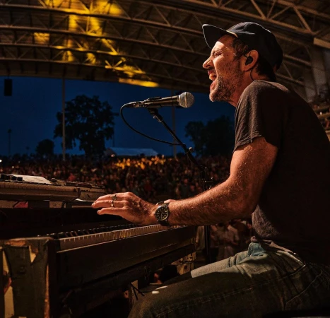 Man passionately singing and playing keyboard at outdoor concert in dark t-shirt, jeans, and ball cap in front of a crowd.
