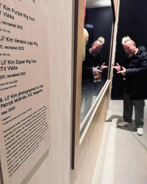 Adult male with bleached hair observes a museum exhibit with Lil' Kim wig display and signage detailing the wigs and photographer.