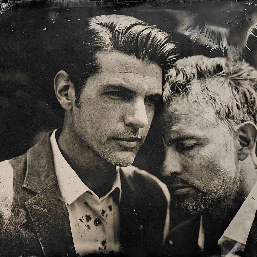 Black and white portrait of two adult men in jackets, heads close together, deep shadows and vintage processing.