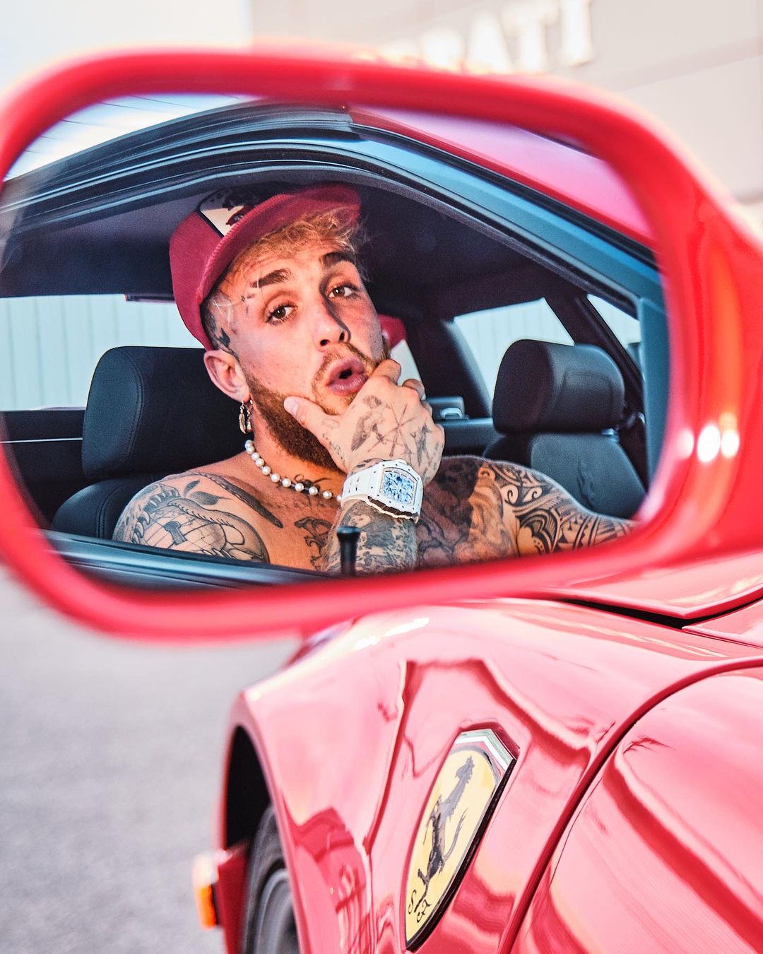 Jake Paul inside a red Ferrari, reflected in the rearview mirror, wearing a red baseball cap, tattoos, pearl necklace, and watch.