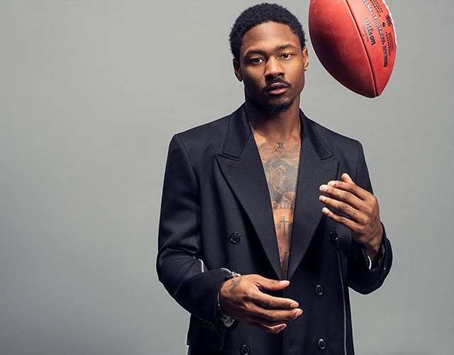 Portrait of a Black male athlete in a black blazer with an American football, set against a grey backdrop in a photo studio.