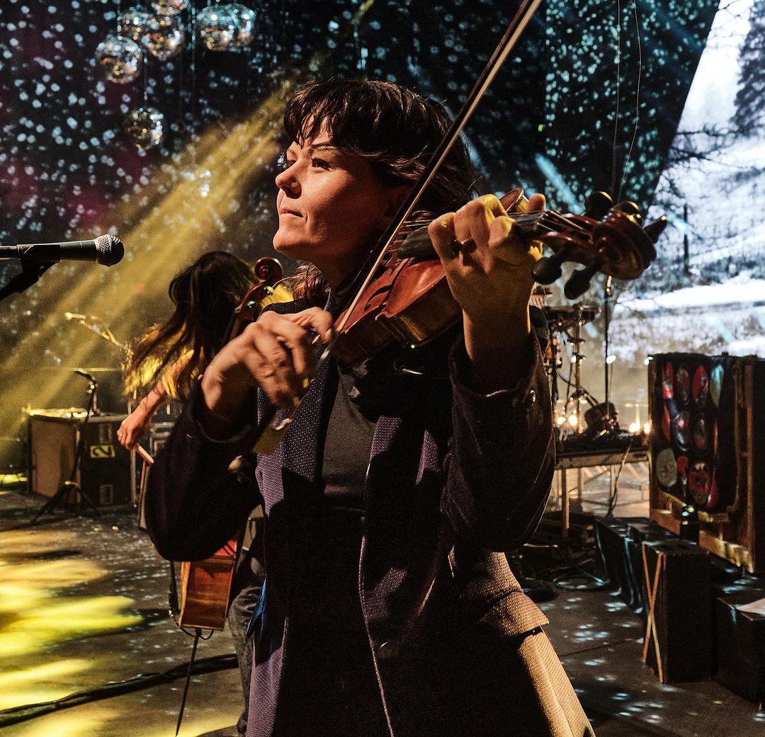 Musician plays violin on stage with orchestra and abstract lighting. Woman in coat performing with stringed instrument in concert hall.