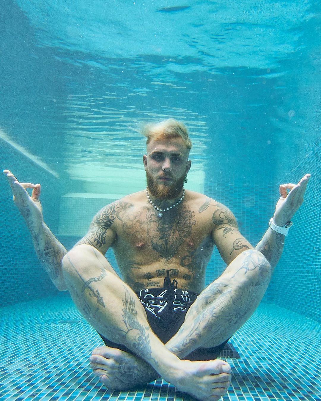 Man with tattoos and blonde hair underwater in a seated lotus pose with his arms raised in a yoga mudra, eyes facing the camera.
