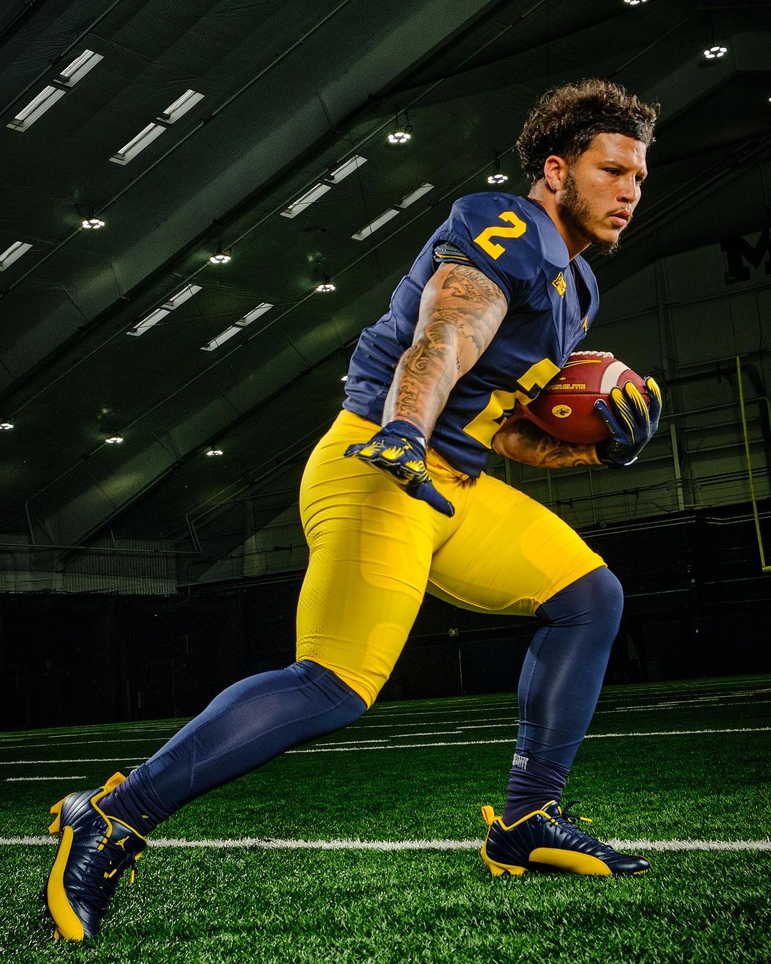 Football player in blue and yellow uniform runs with the ball on green turf inside a stadium with lights and a dark ceiling.
